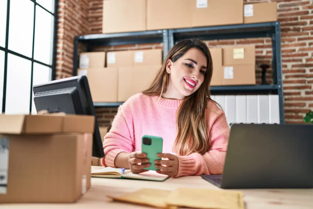 Young Beautiful Hispanic Woman Ecommerce Business Worker Using Smartphone Office - Sgariboldi Consulting Business